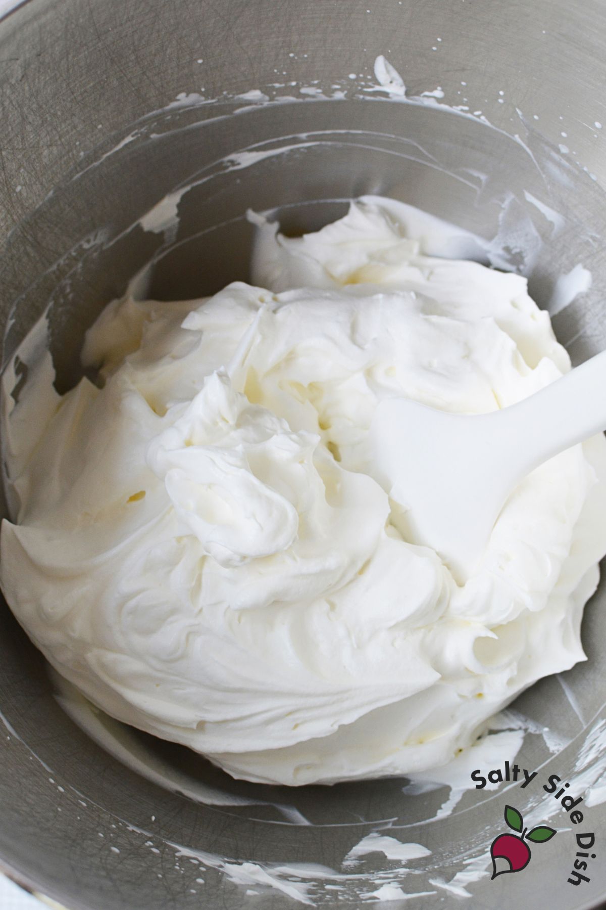 Thick homemade whipped cream in a stainless steel mixing bowl with a white spatula.